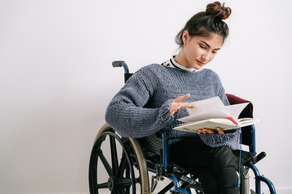 Woman in a blue knitted sweater sits in a wheelchair and reads an open book.